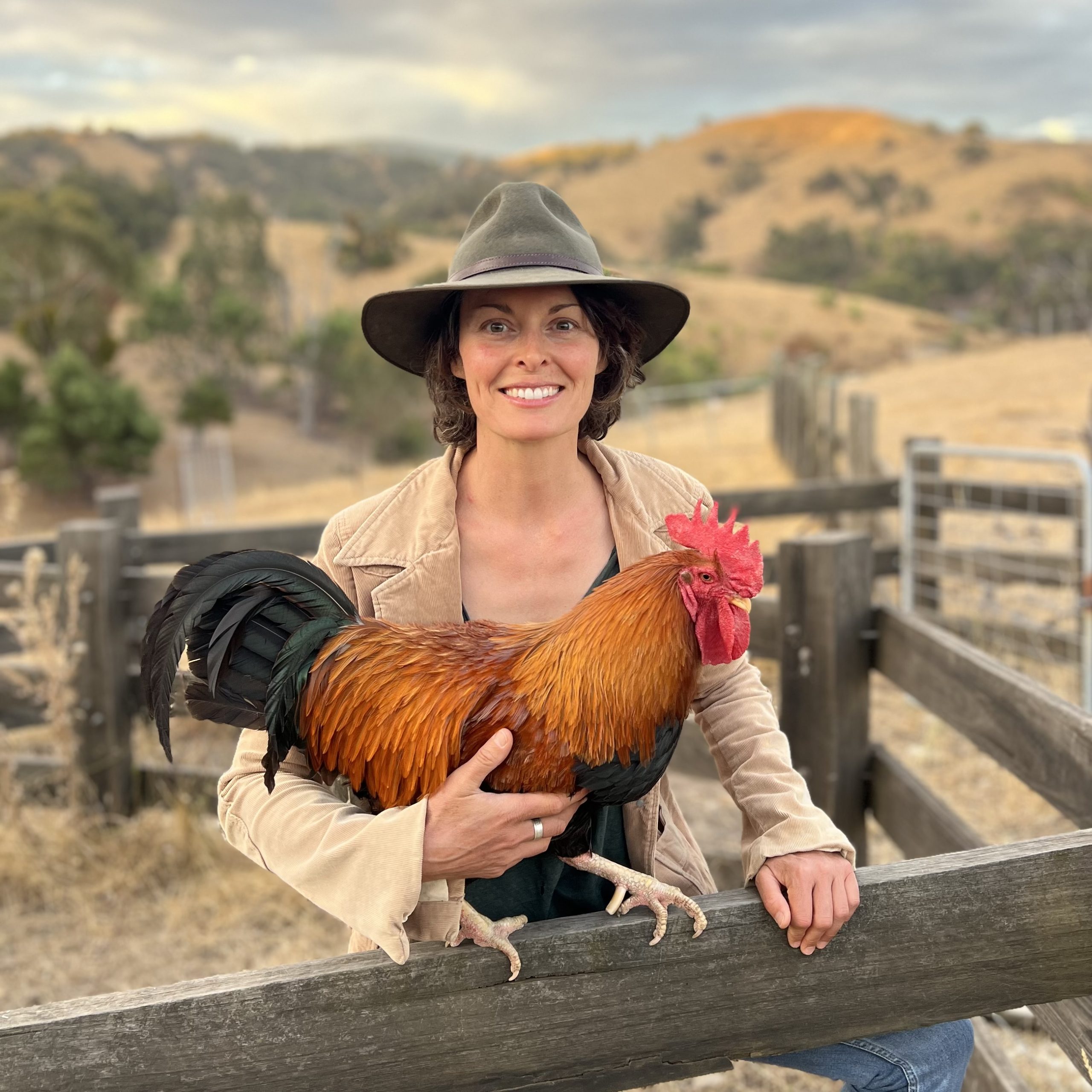 Heritage chicken breeder Jane Cowan leans on a wooden fence with her Welsummer rooster, Tabasco, wearing an olive green Akubra hat and cord jacket which matches the tones of the sunburnt hills in the background.